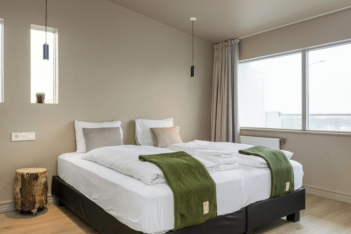 Una cama bien hecha con sábanas blancas y un plaid verde, un taburete de madera y una ventana con cortinas.
