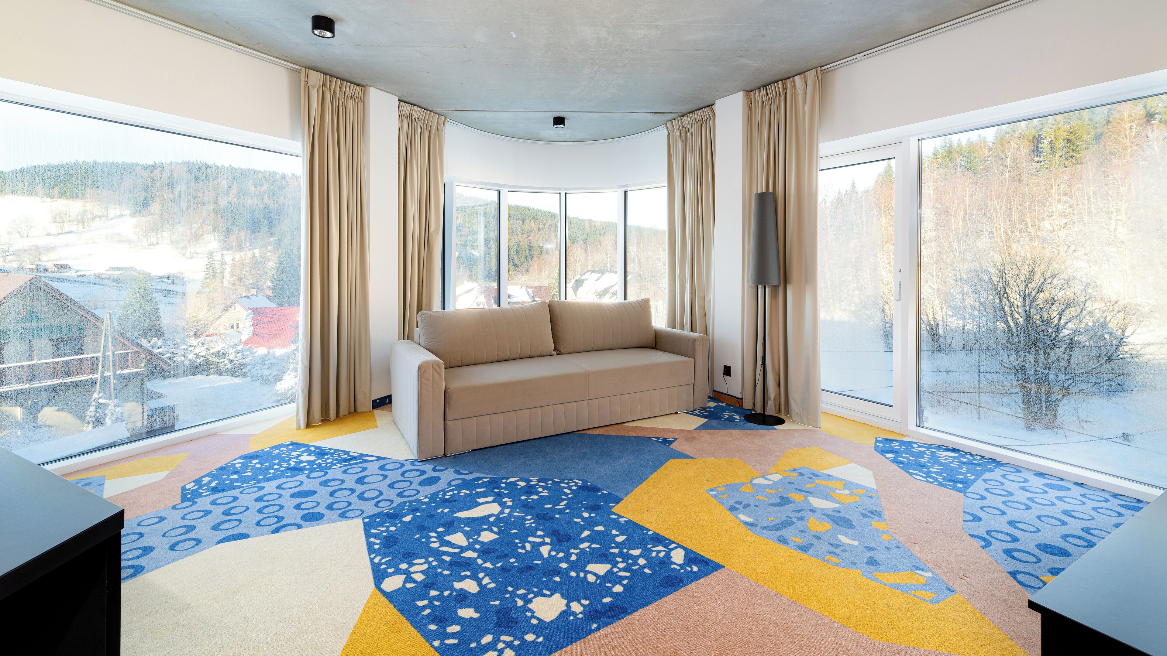 A modern living room with a beige sofa, large windows, and a colorful geometric carpet.