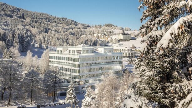 Un hotel moderno con varios pisos, rodeado de árboles nevados y montañas al fondo.