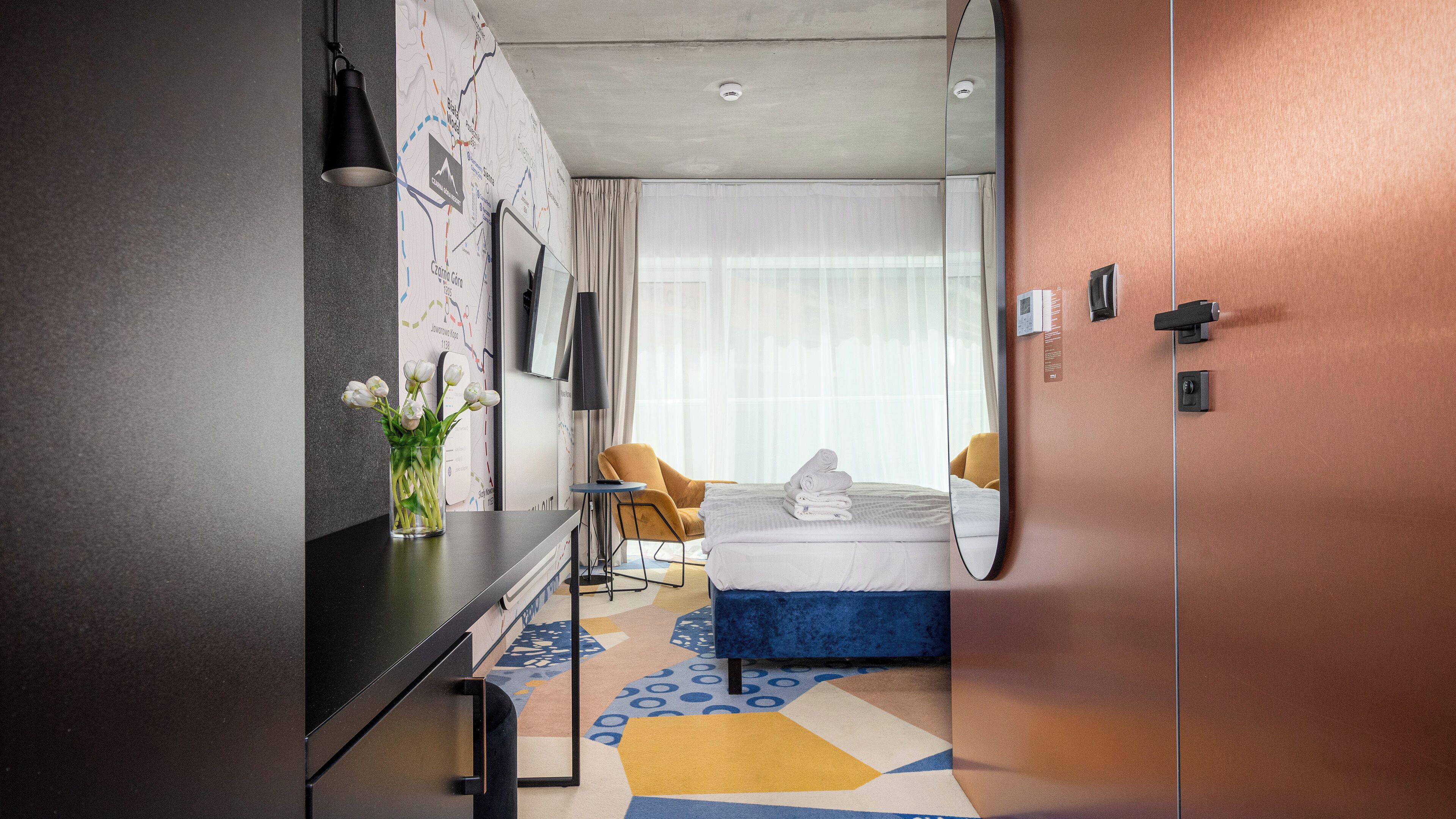 A modern hotel room with a bed, a desk, a chair, and a large window.