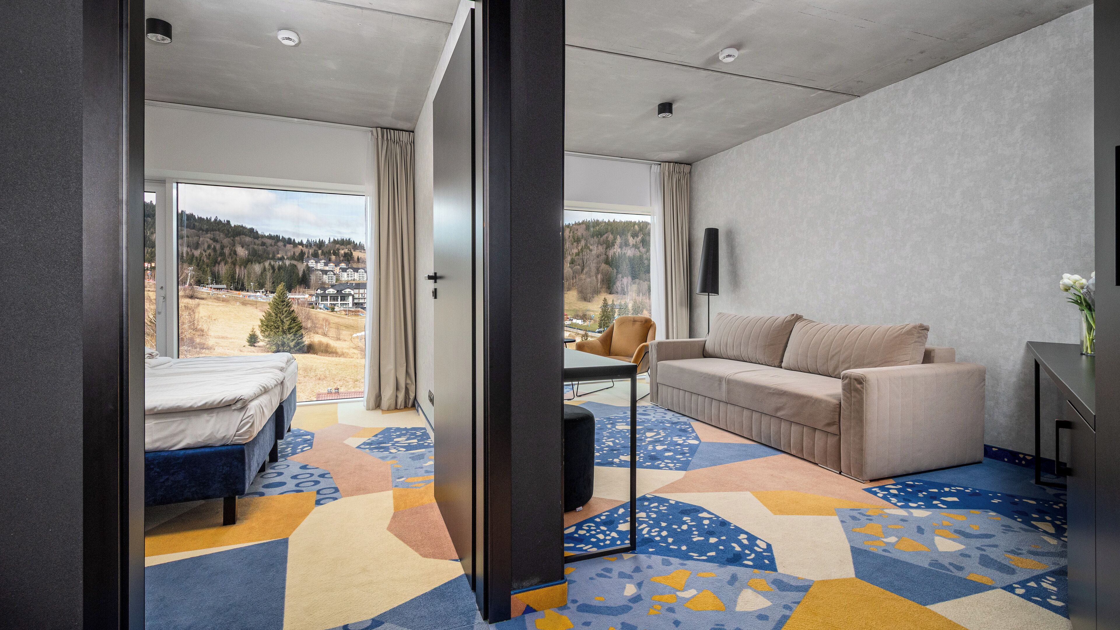 A modern hotel room with a bed, a sofa, a coffee table, and a view of the outdoors.