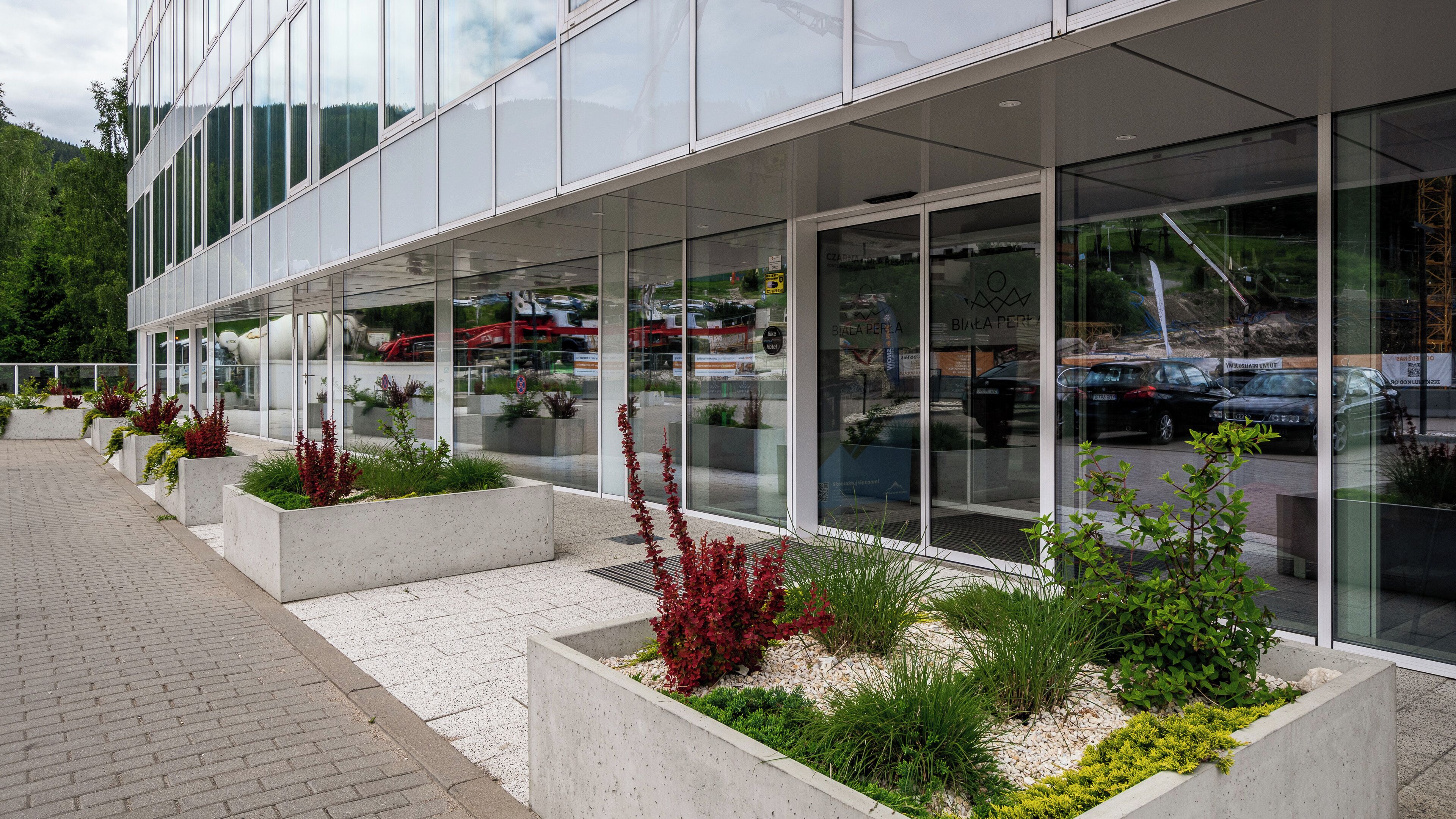 A modern building with large glass windows, outdoor seating areas with planters, and a paved walkway.
