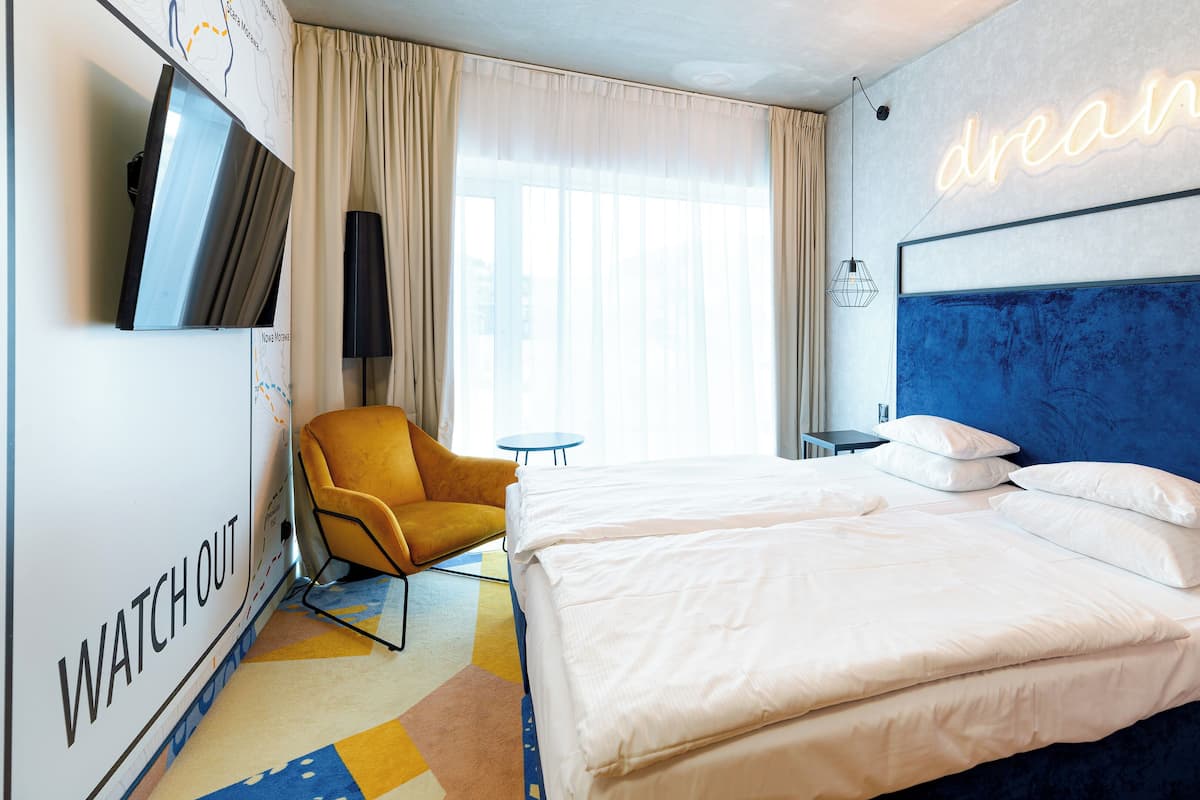 Une chambre d’hôtel avec un grand lit, un fauteuil jaune, une petite table et un néon représentant le mot « rêve » accroché au mur.
