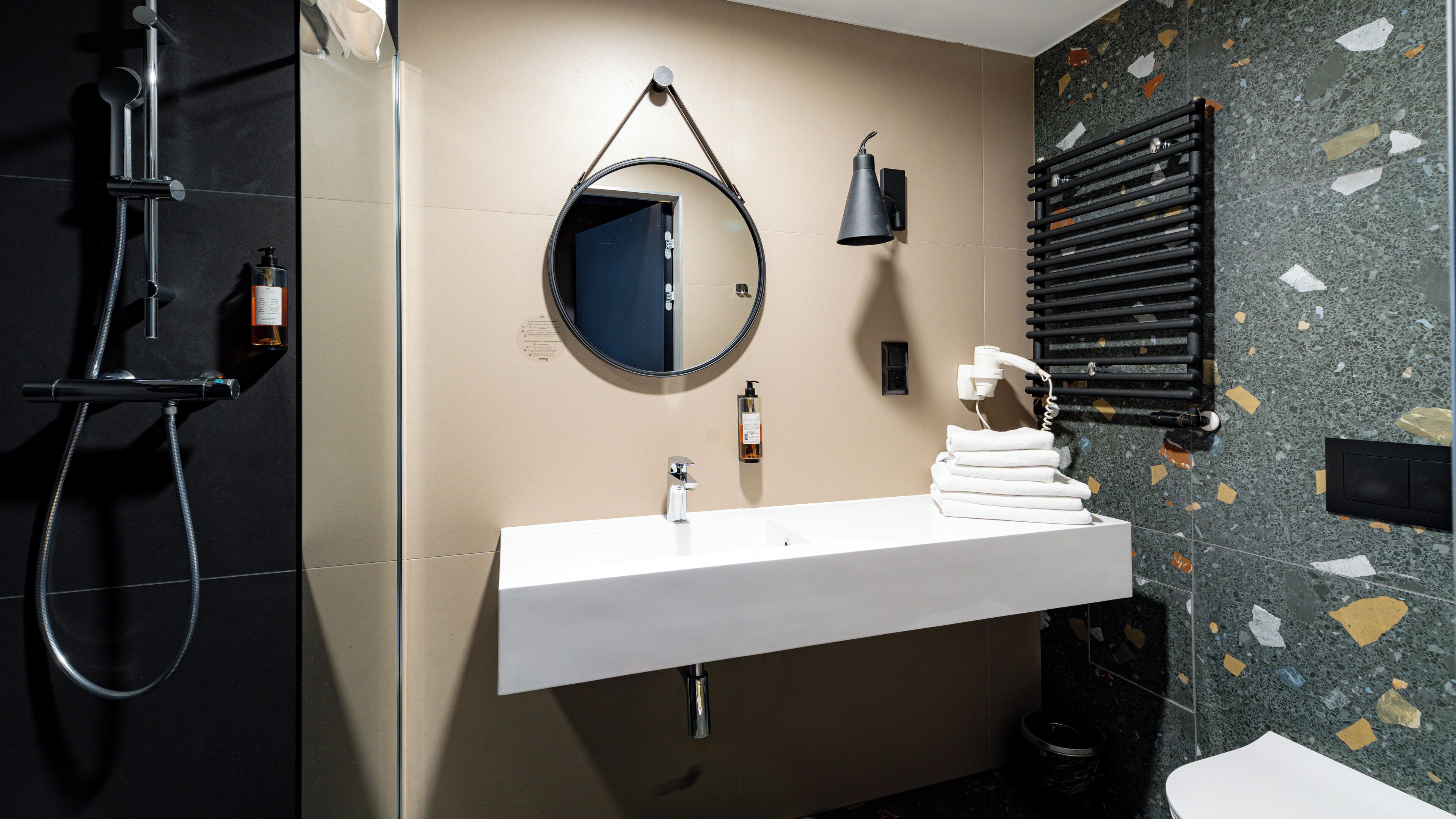 Un baño moderno con un lavabo blanco, un espejo redondo, un cabezal de ducha de mano y un perchero para toallas.