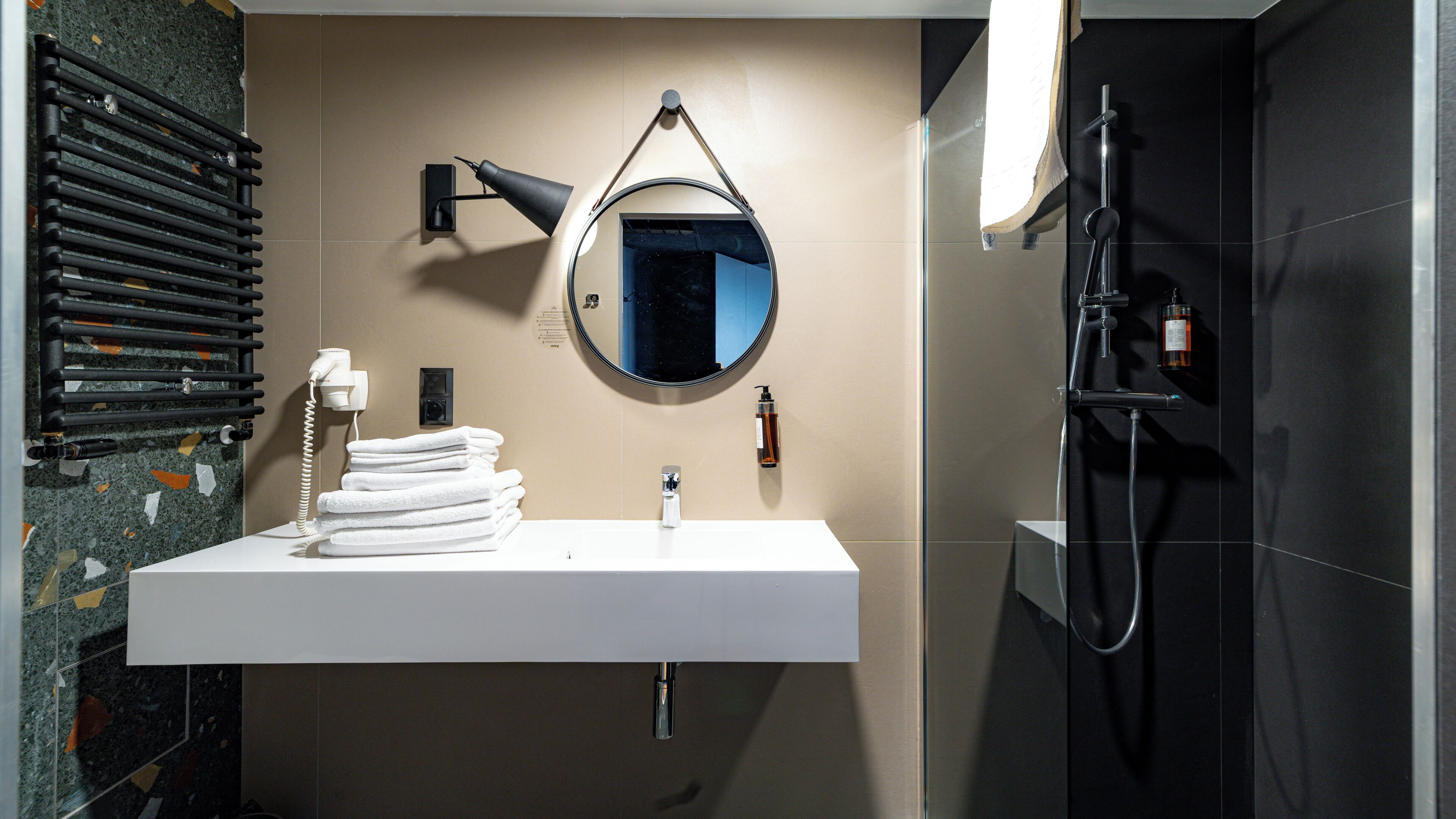 A bathroom with a white sink, a round mirror, a towel rack, a handheld showerhead, and a towel dispenser.