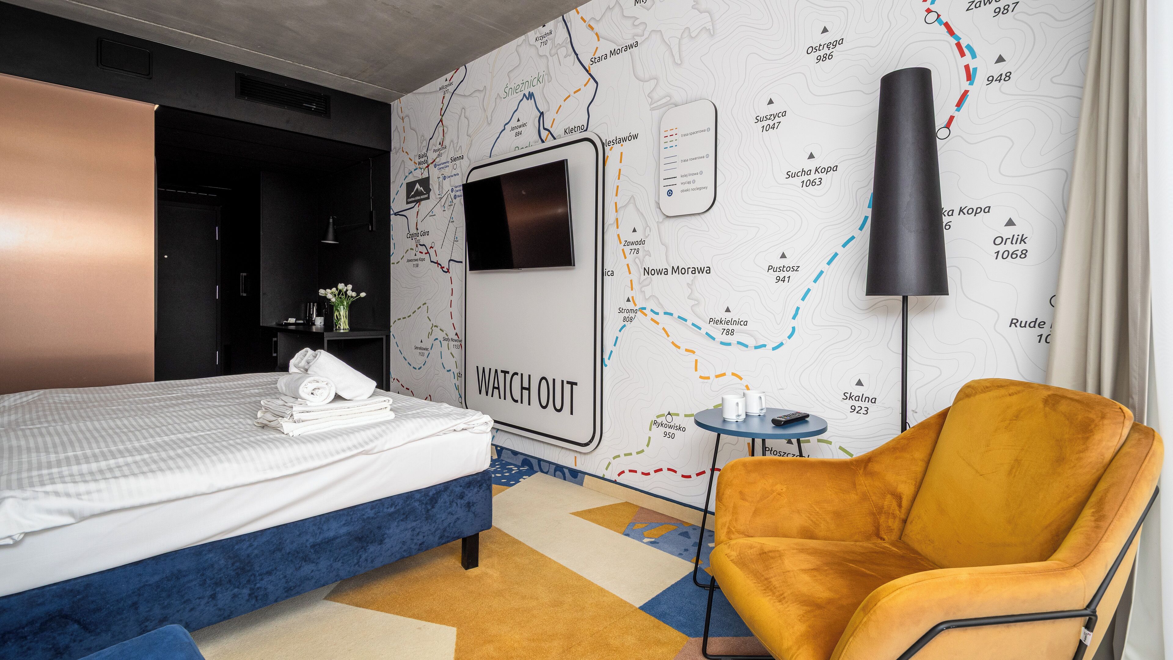 A hotel room with a bed, a map wall, a TV, a small table, and a yellow armchair.