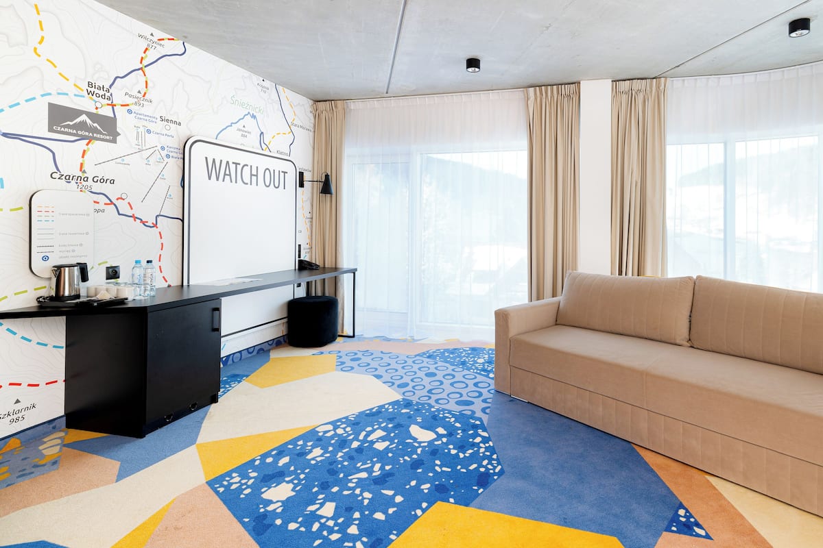 Une chambre d’hôtel moderne avec une grande fenêtre, un canapé, un bureau avec une carte et un panneau indiquant « ATTENTION ».