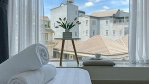 A hotel room with a neatly made bed, a small table with a plant, and a view of buildings through large windows. - Lam Anh Hotel (Xuan Huong - Da Lat)