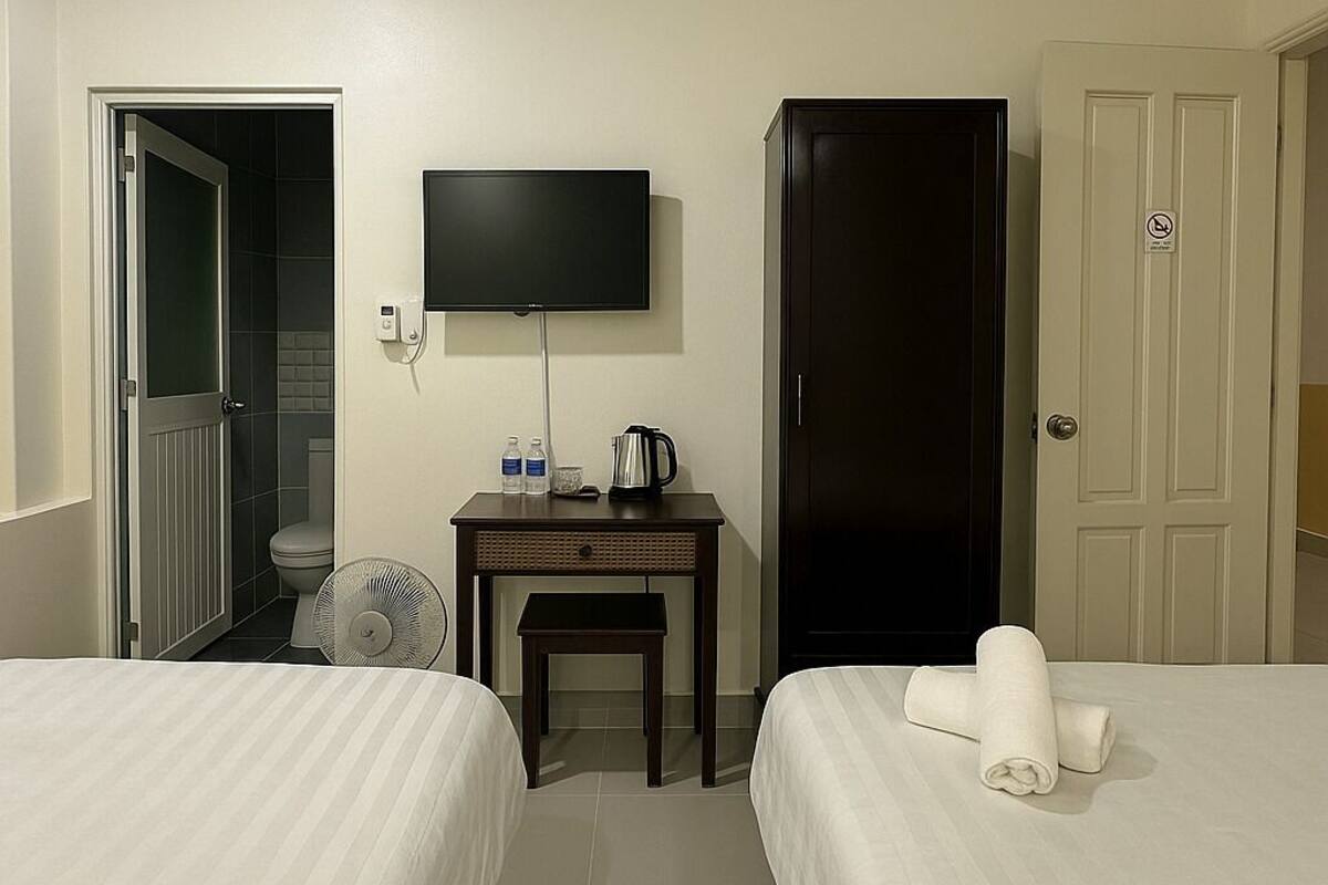 A hotel room with two beds, a TV, a desk with a kettle, a fan, and a wardrobe.
