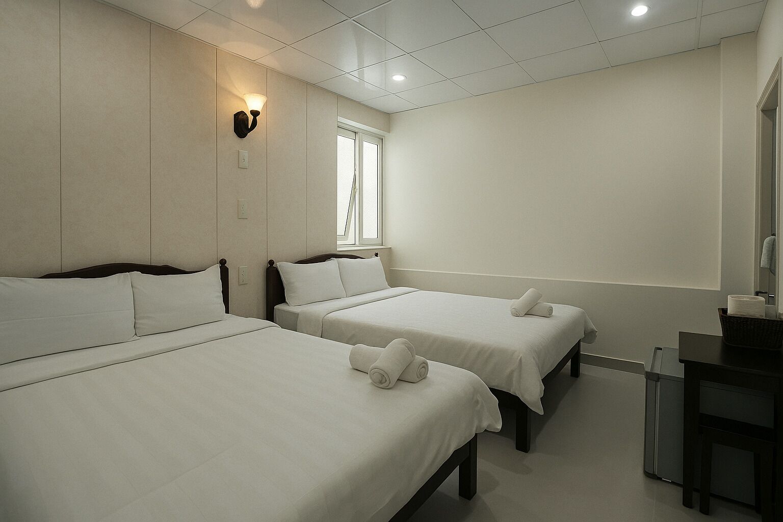 A hotel room with three single beds, a small desk, a window, and a wall-mounted light fixture.