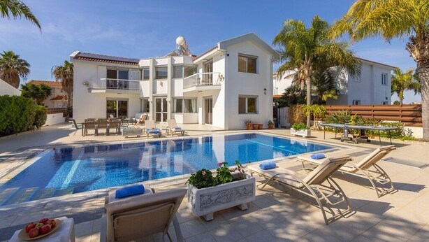 A two-story white building with a swimming pool, lounge chairs, and palm trees.