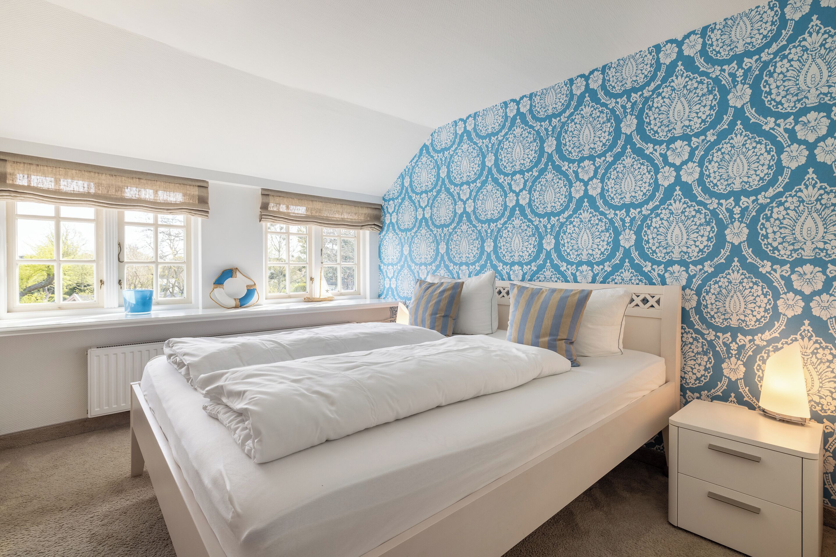 Une chambre avec deux lits, une fenêtre donnant sur un beau paysage, une table de chevet avec une lampe et un mur décoré d’un motif.