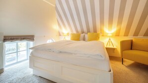 Une chambre avec un grand lit, une fenêtre, un fauteuil jaune et des murs à rayures.