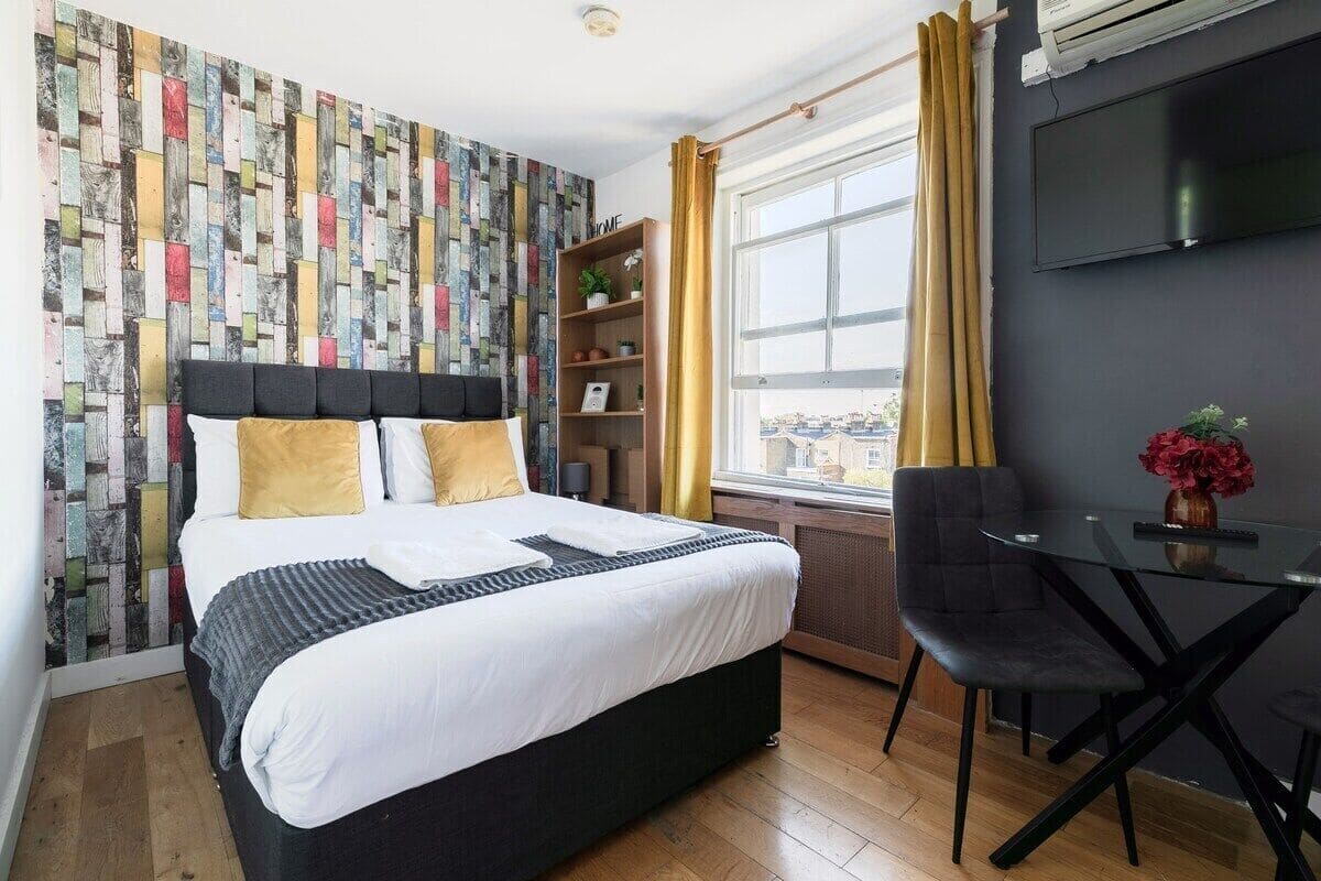 A modern hotel room with a large bed, a small table, a chair, a TV, and a window with curtains.
