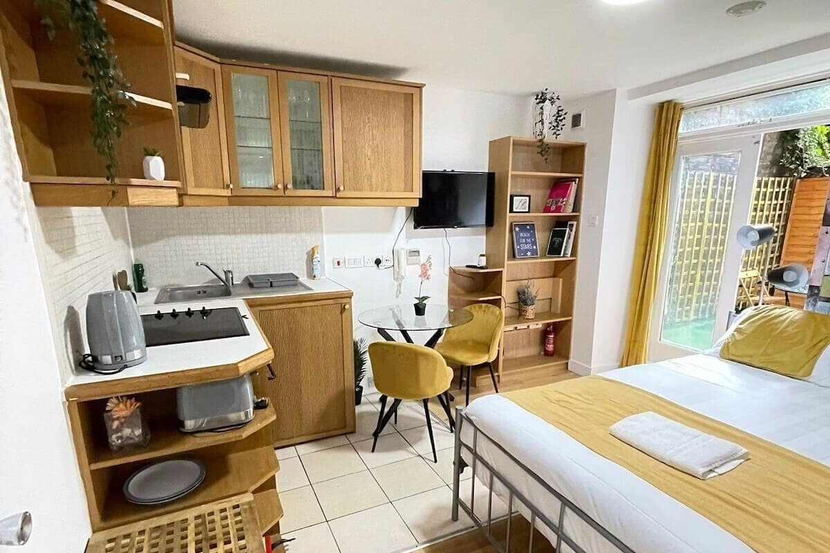 A compact hotel room with a kitchenette, a bed with a wooden headboard, a small desk, and a chair.