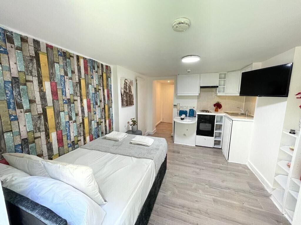 A compact bedroom with a bed, a wall-mounted TV, and a kitchenette with white cabinets and a microwave.