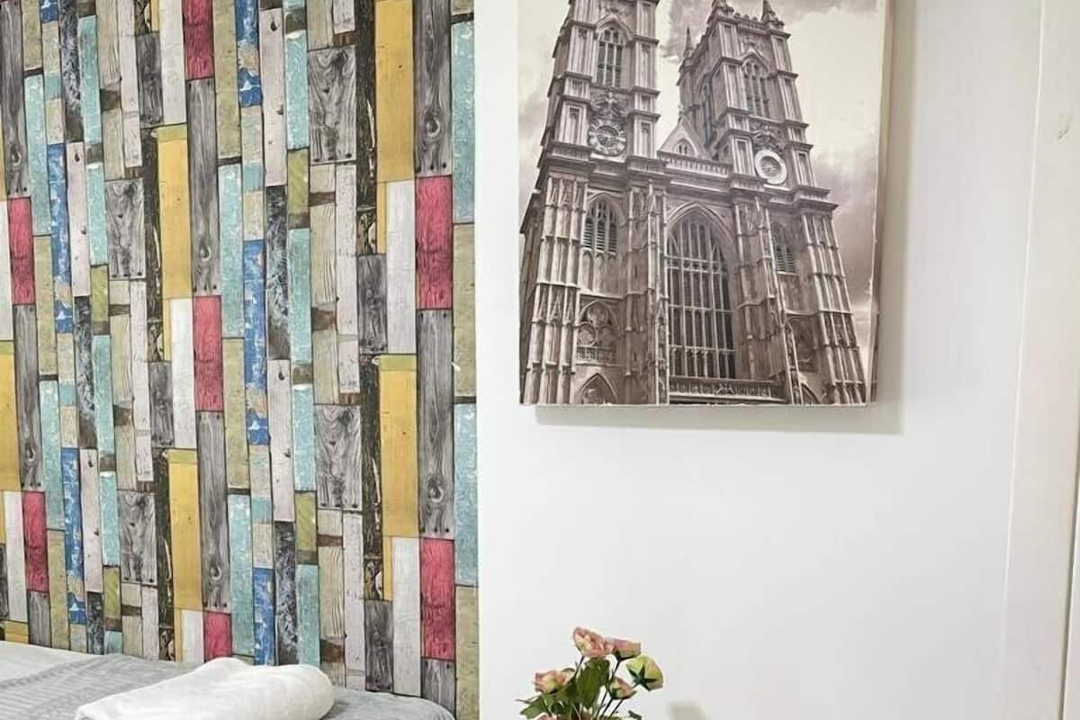 A bedroom with a bed, a colorful wooden wall panel, a framed picture of a building, and a small table with a vase of flowers.