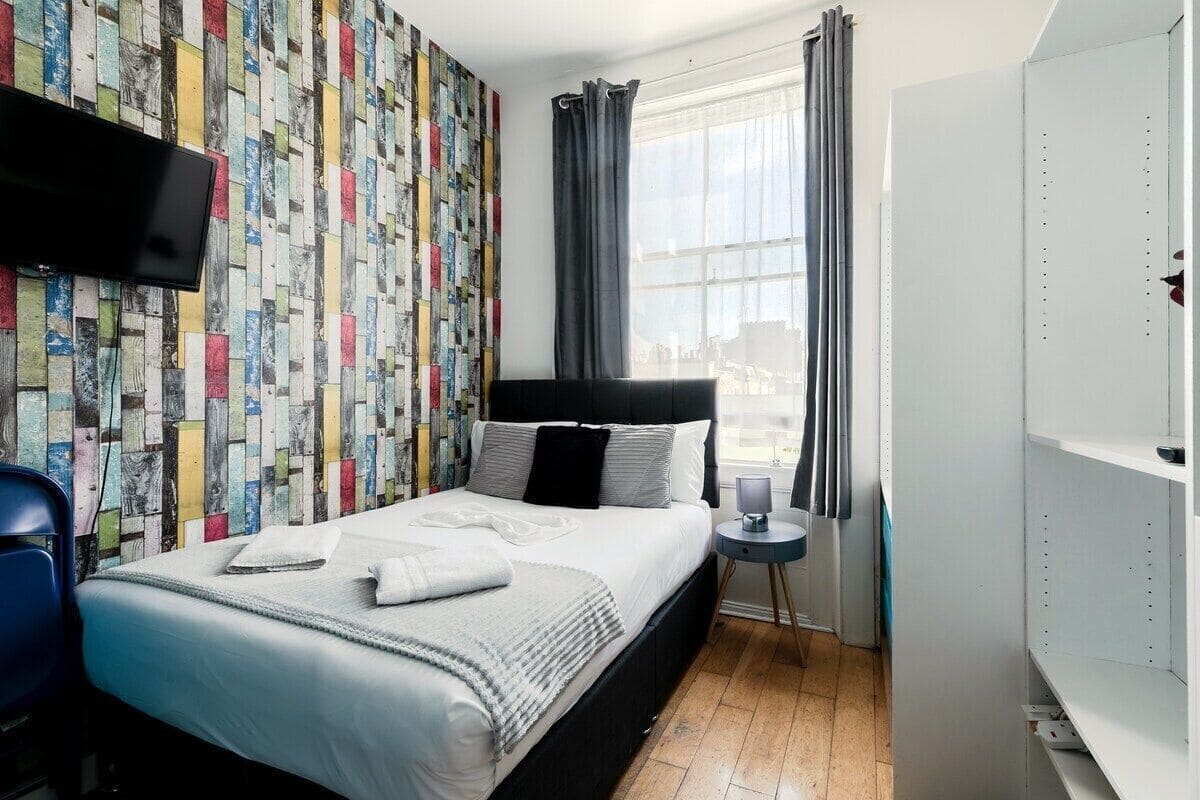 A neatly made bed with pillows and a blanket, a bedside table with a lamp, a TV mounted on the wall, and a window with curtains.