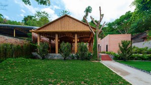 A wooden pavilion with a thatched roof, surrounded by a well-maintained lawn and a brick building. - Phuc Thang Resort  (Phu Quoc)