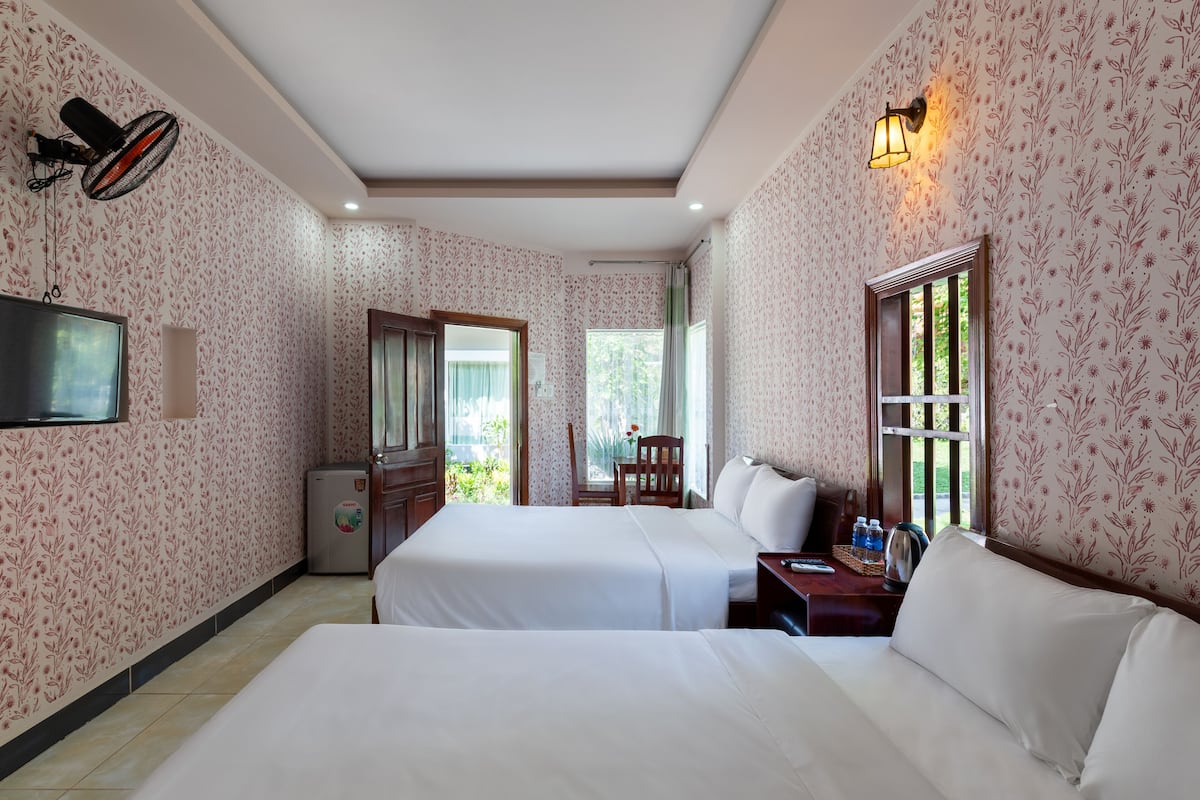 A hotel room with two beds, a TV, a fan, a desk with a lamp, and a window with a view of greenery.