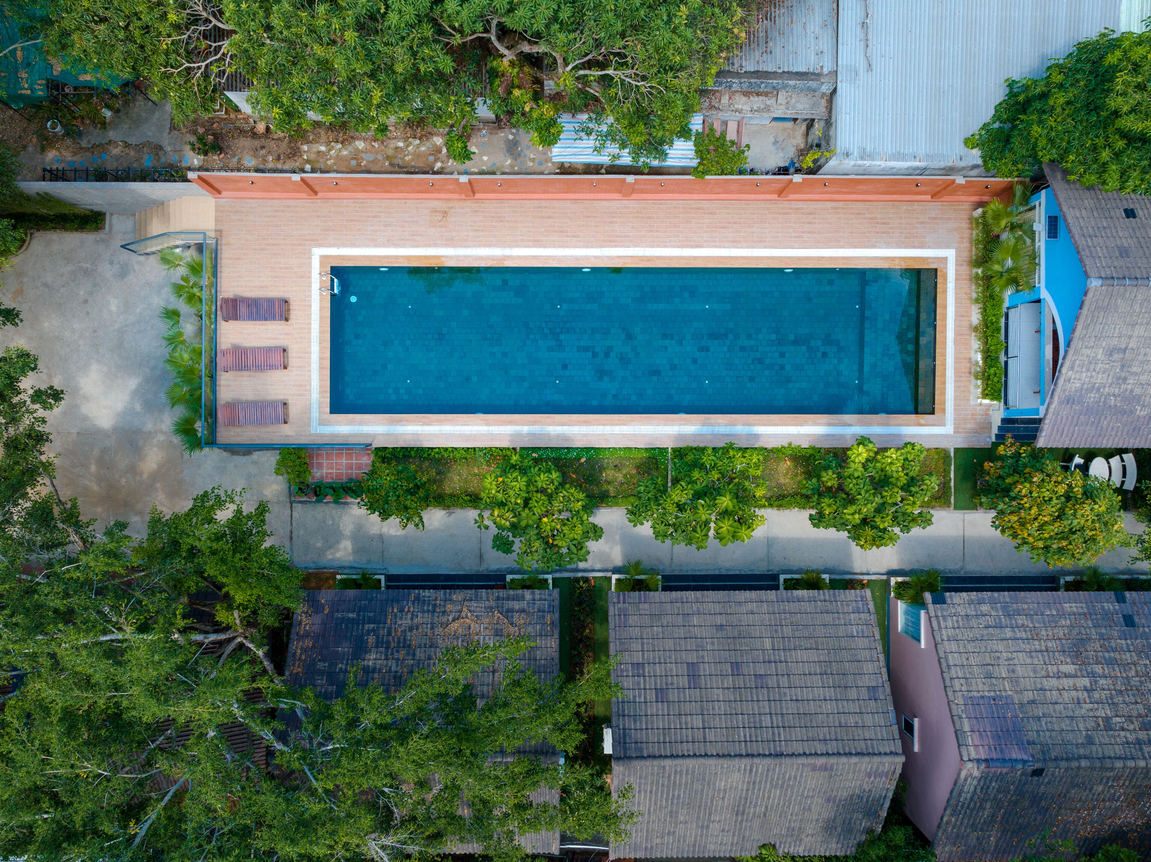An aerial view of a rectangular swimming pool surrounded by lush greenery, with adjacent buildings and a clear sky.