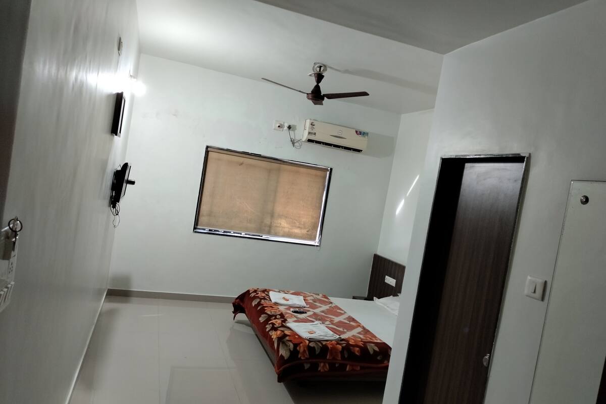 A small, sparsely furnished room with a bed, a window, an air conditioning unit, and a ceiling fan.