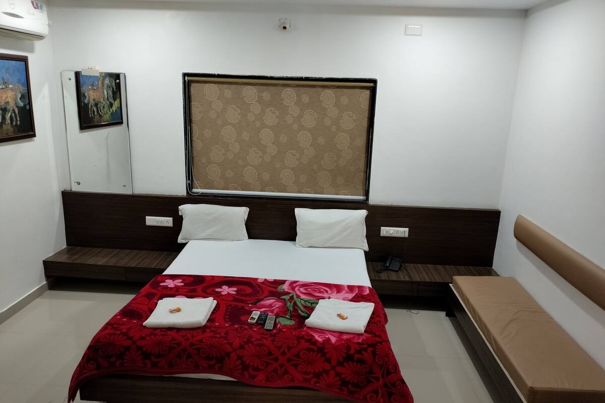A hotel room with a bed, two pillows, a bedside table, a telephone, and a window with a patterned curtain.