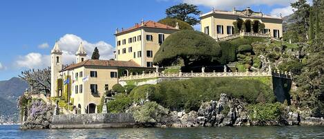 A large, multi-story building with a tower, situated on a lakeside with a well-maintained garden and a stone wall.