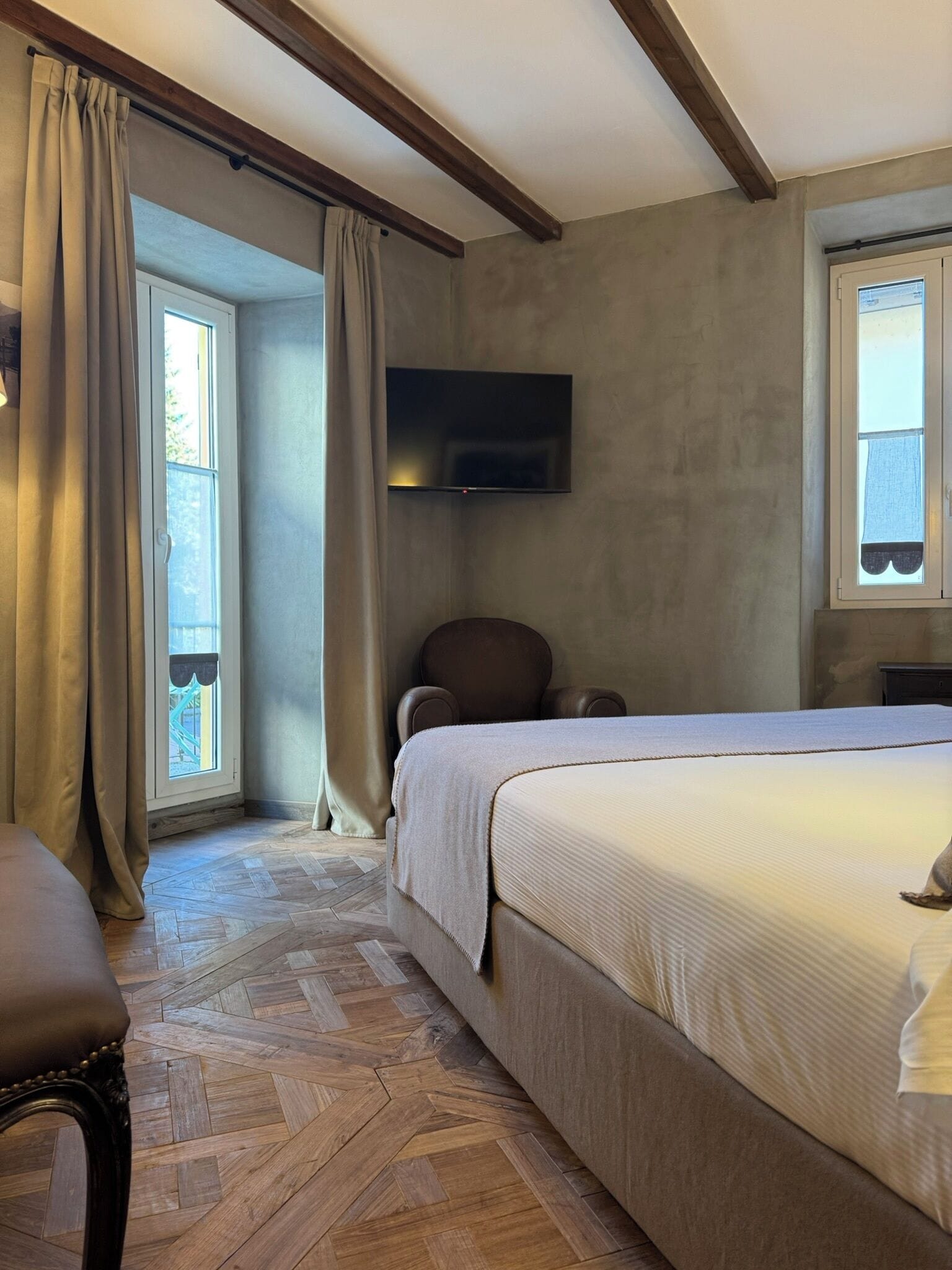 Un dormitorio con una cama grande, una silla, un televisor montado en la pared y una ventana con cortinas.