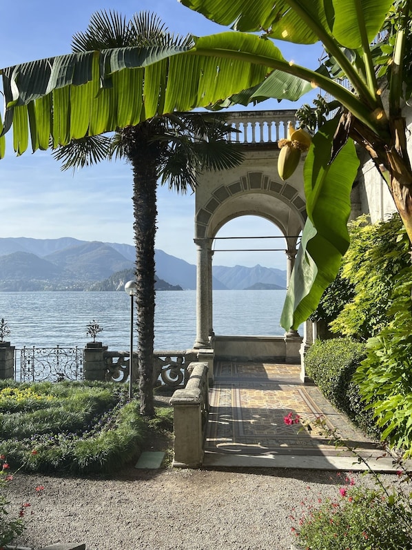 A garden pathway leading to a view of a lake with mountains in the background, surrounded by lush greenery and a decorative archway.