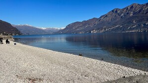 A pebbled beach with a calm lake, mountains in the background, and a clear blue sky. - Al Molo 5 Apartments (Oliveto Lario)