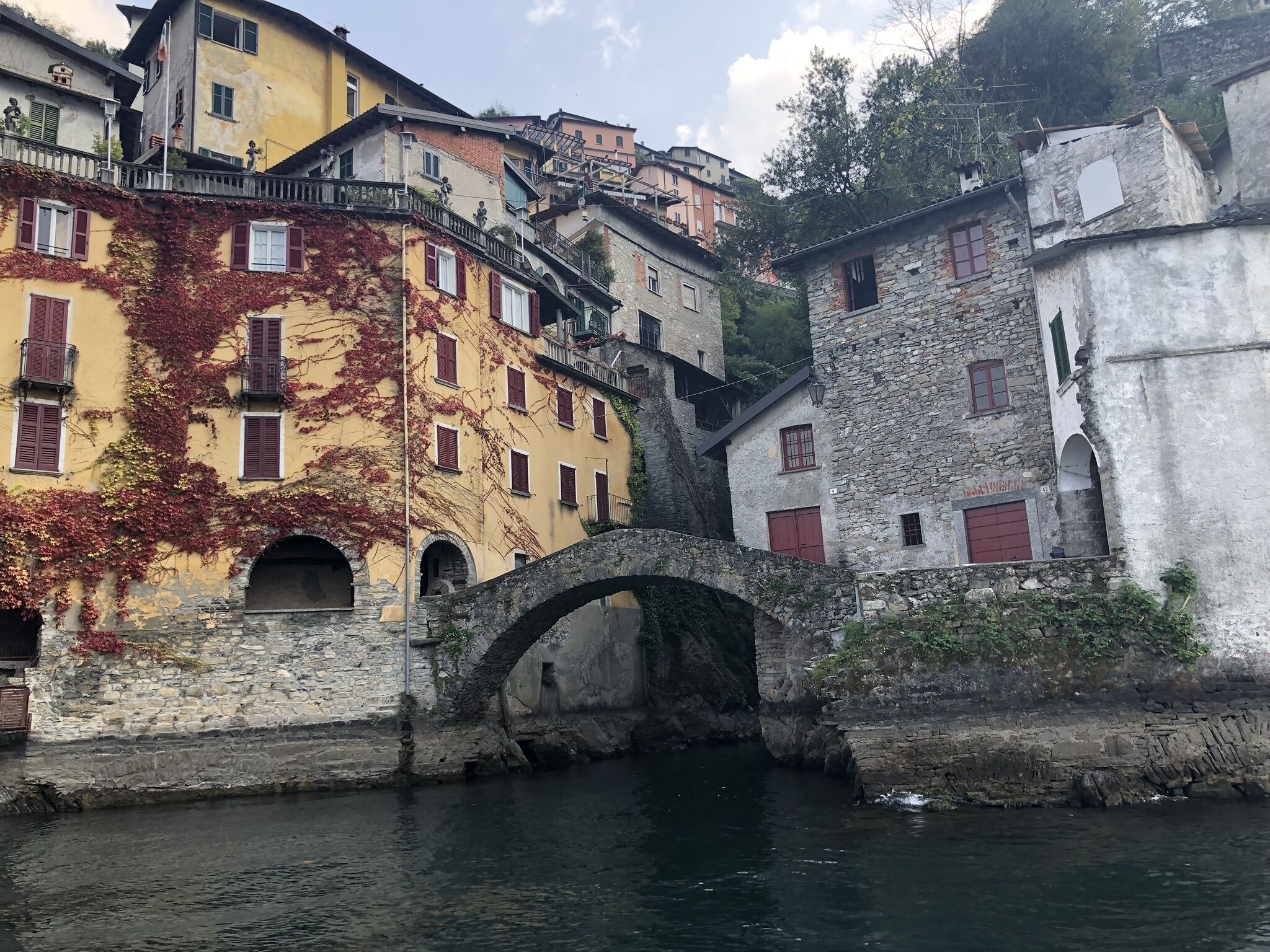 Un pont de pierre enjambe une rivière et relie des bâtiments dotés d’arcs en plein cintre et de volets rouges.