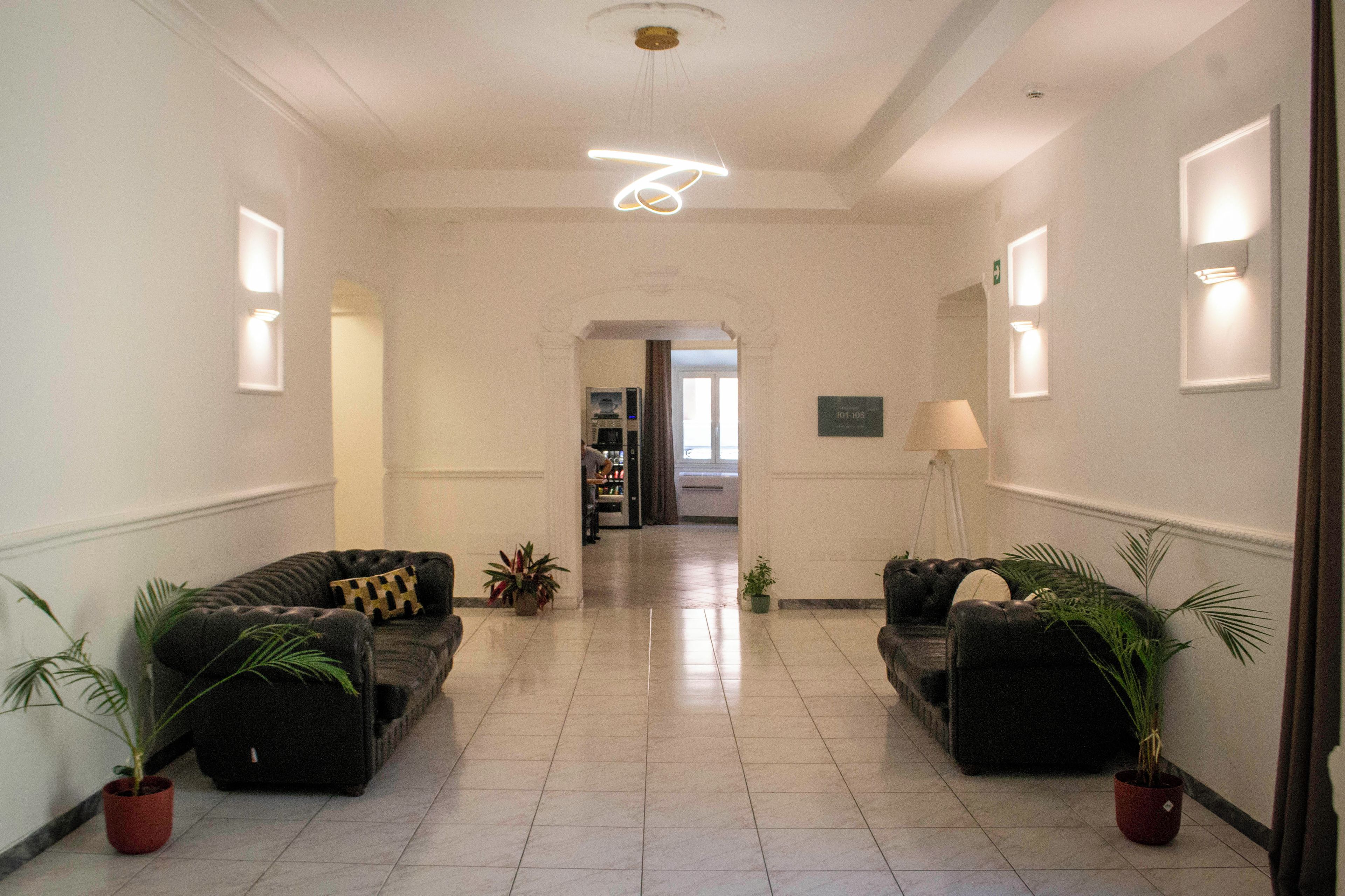 Una hall d'albergo con un corridoio centrale, due divani con piante in vaso, una macchina del caffè e una lampada.