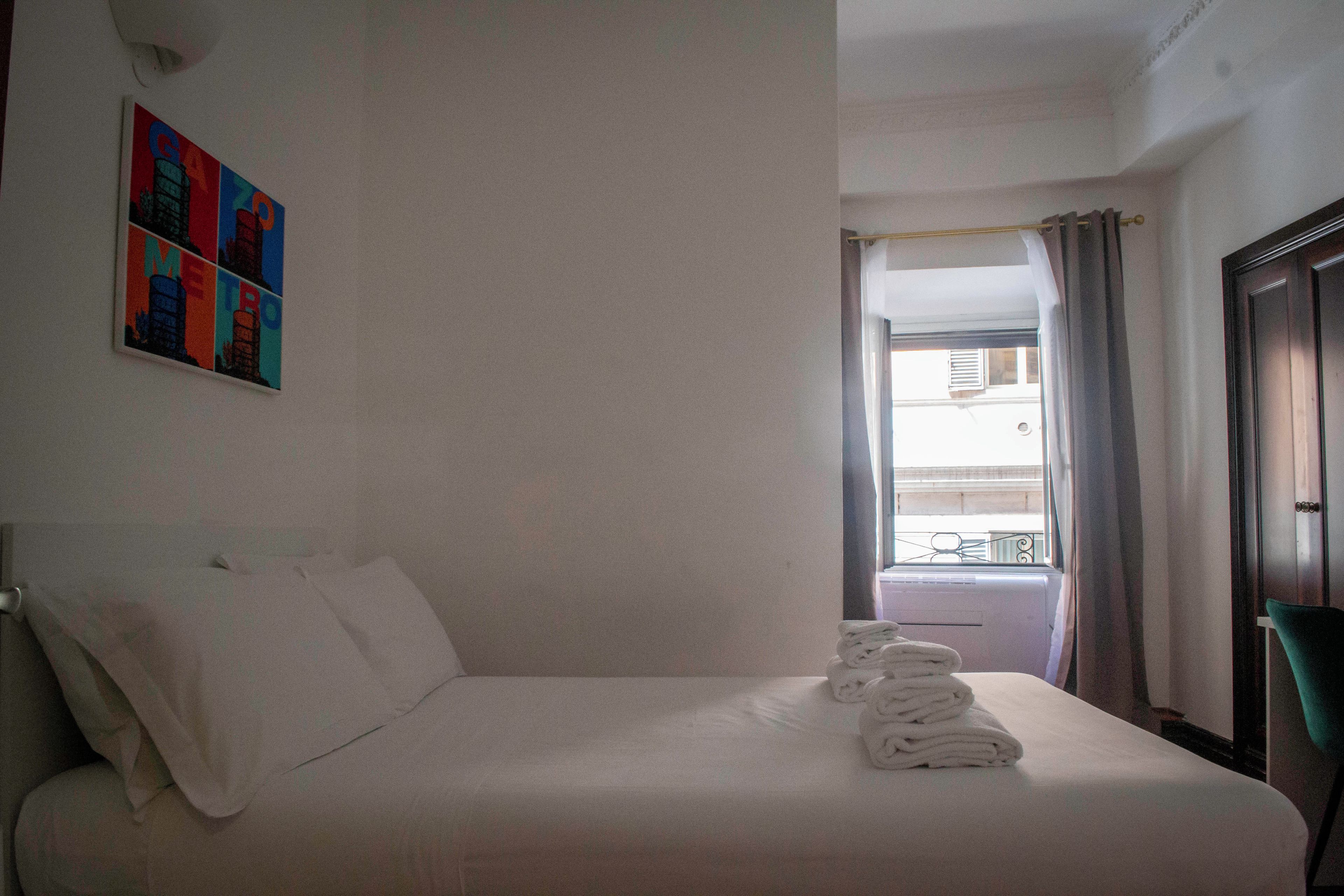 A hotel room with a bed, pillows, a folded towel, a window with curtains, and a painting on the wall.