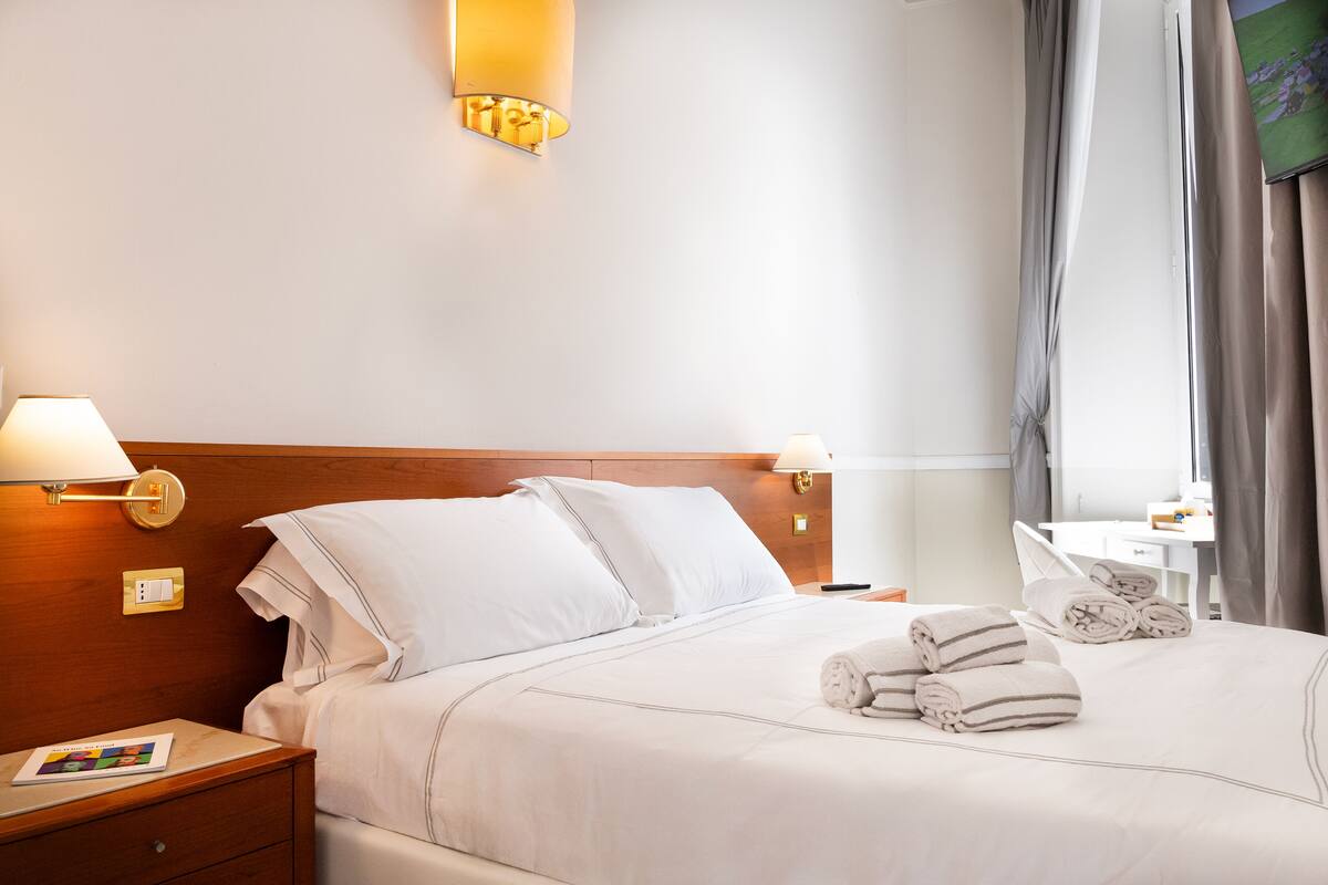 A neatly made bed with white linens, two bedside lamps, a small bedside table with a book, and a window with curtains.