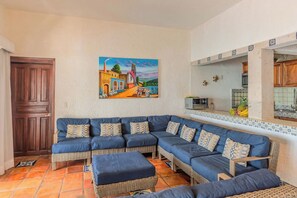 Una sala de estar con un sofá seccional, un cuadro de un pueblo y una cocina con salpicadero de azulejos.