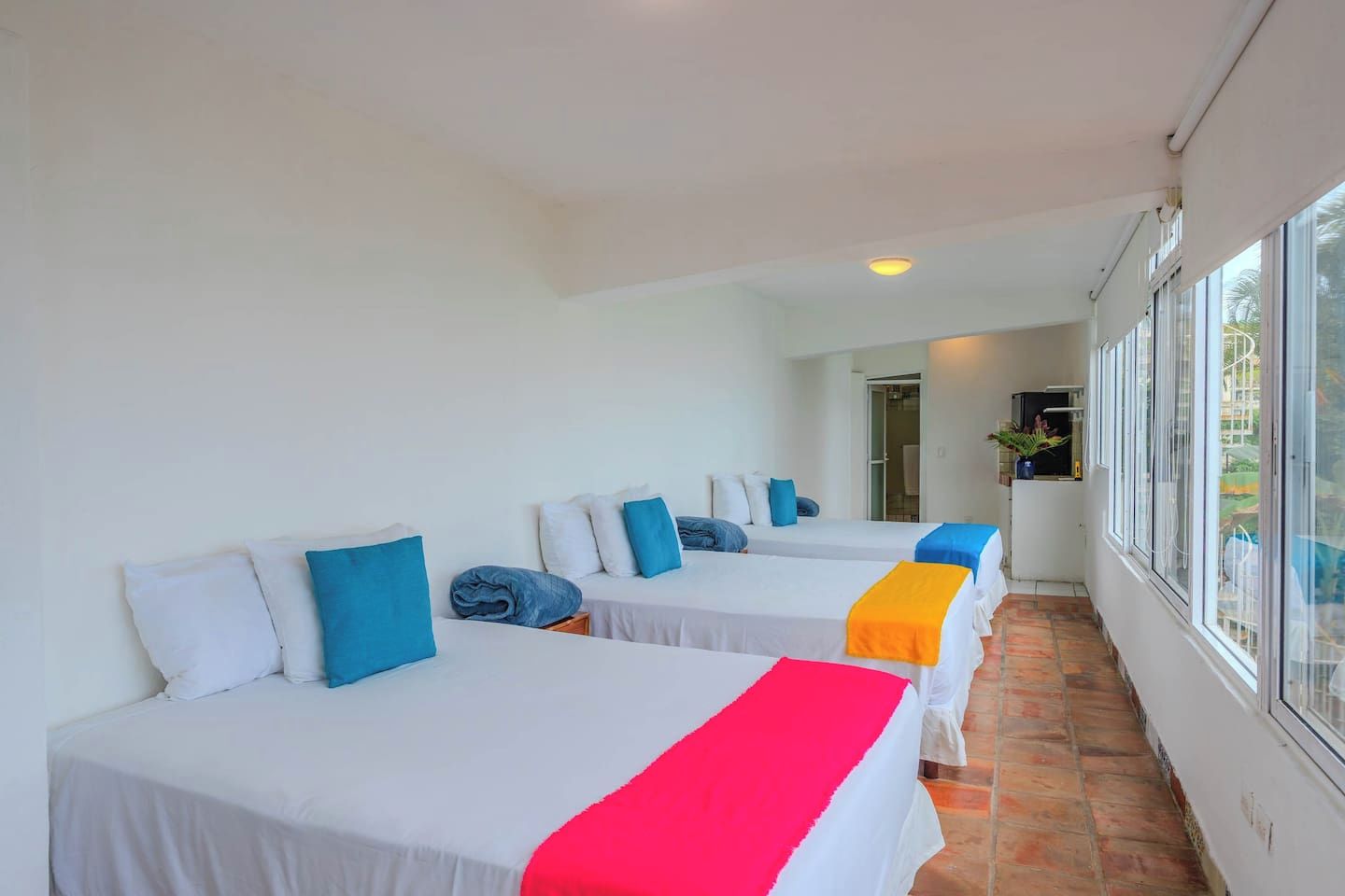A corridor with three beds, each with a white bedspread and blue pillows, leading to a room with a window.