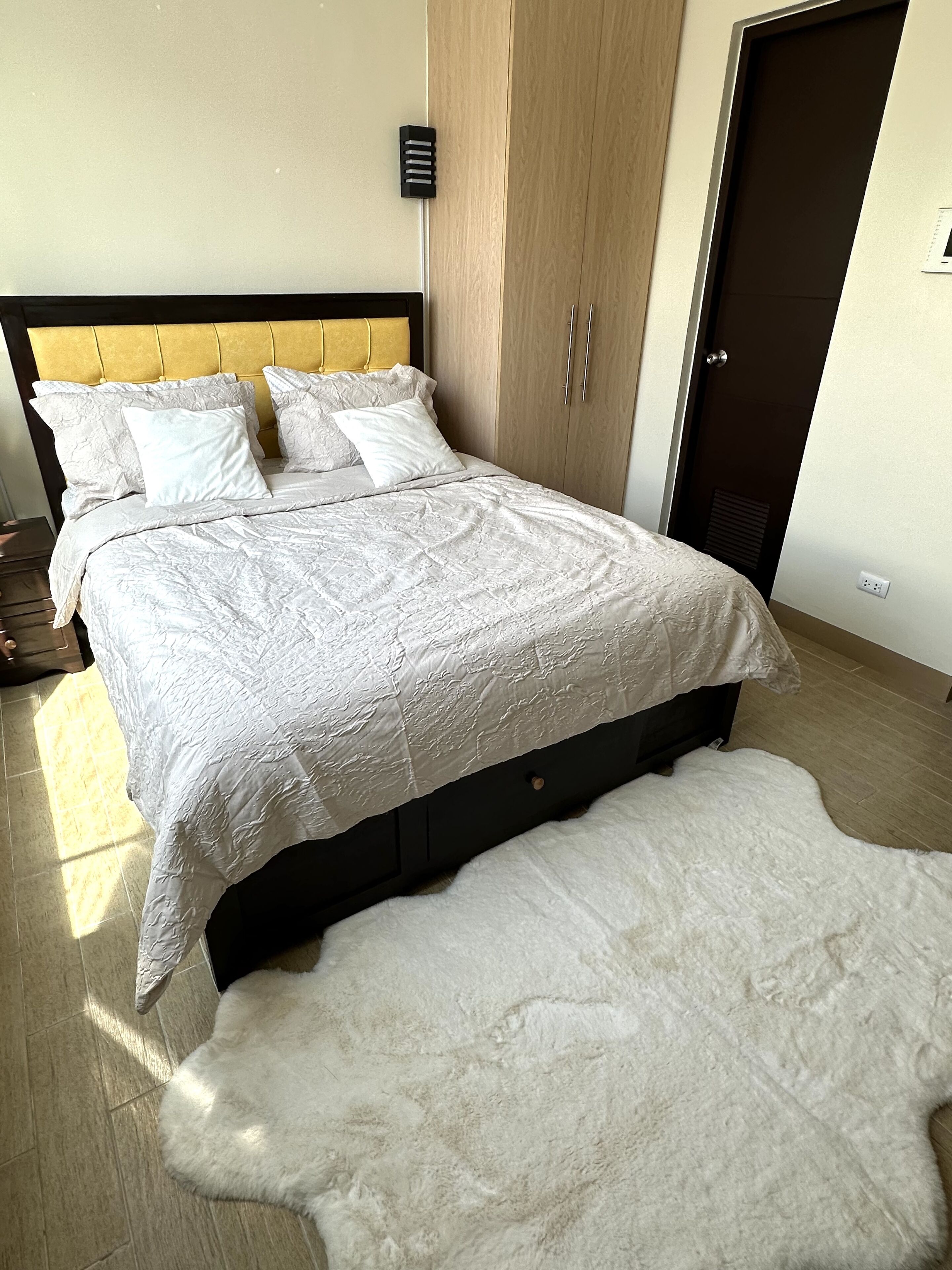 Un letto rifatto con un piumino bianco e cuscini, un armadio in legno e un tappeto morbido e bianco sul pavimento.