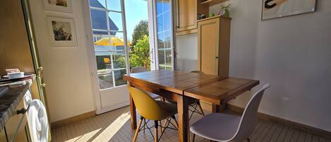 Uma área de refeições com uma mesa de madeira e duas cadeiras, uma janela com vista para o exterior, e uma porta que dá para outro espaço.