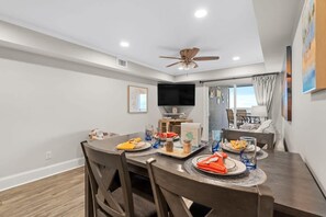 A dining area with a round table, chairs, a ceiling fan, and a television on the wall. - Windy Shores Unit 205 - Oceanfront (North Myrtle Beach)