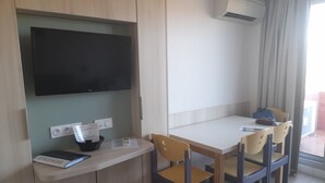 Una habitación pequeña y moderna con una televisión de pantalla plana en la pared, un escritorio con una silla y una mesa con sillas. Hay un aire acondicionado en la pared y una ventana con cortinas.