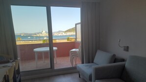 Habitación con sofá, una mesita y una silla. Posee una puerta corrediza de vidrio que da a un balcón con vistas al mar y edificios.