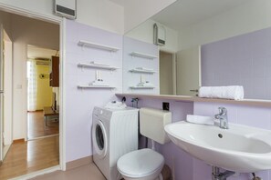 Et moderne badeværelse med vaskemaskine, toilet, håndvask og hylder til håndklæder.