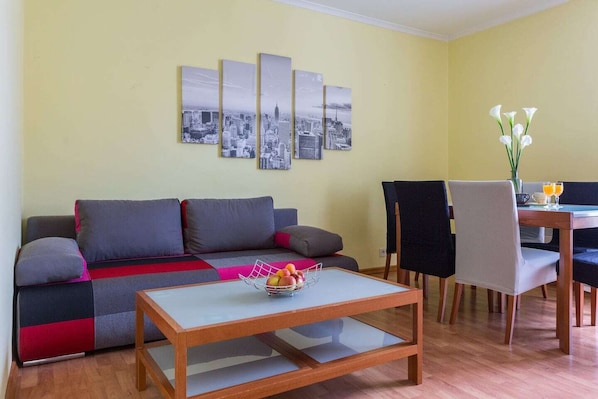 A living room with a sofa, a coffee table with fruit, a dining table with chairs, and a cityscape artwork on the wall.