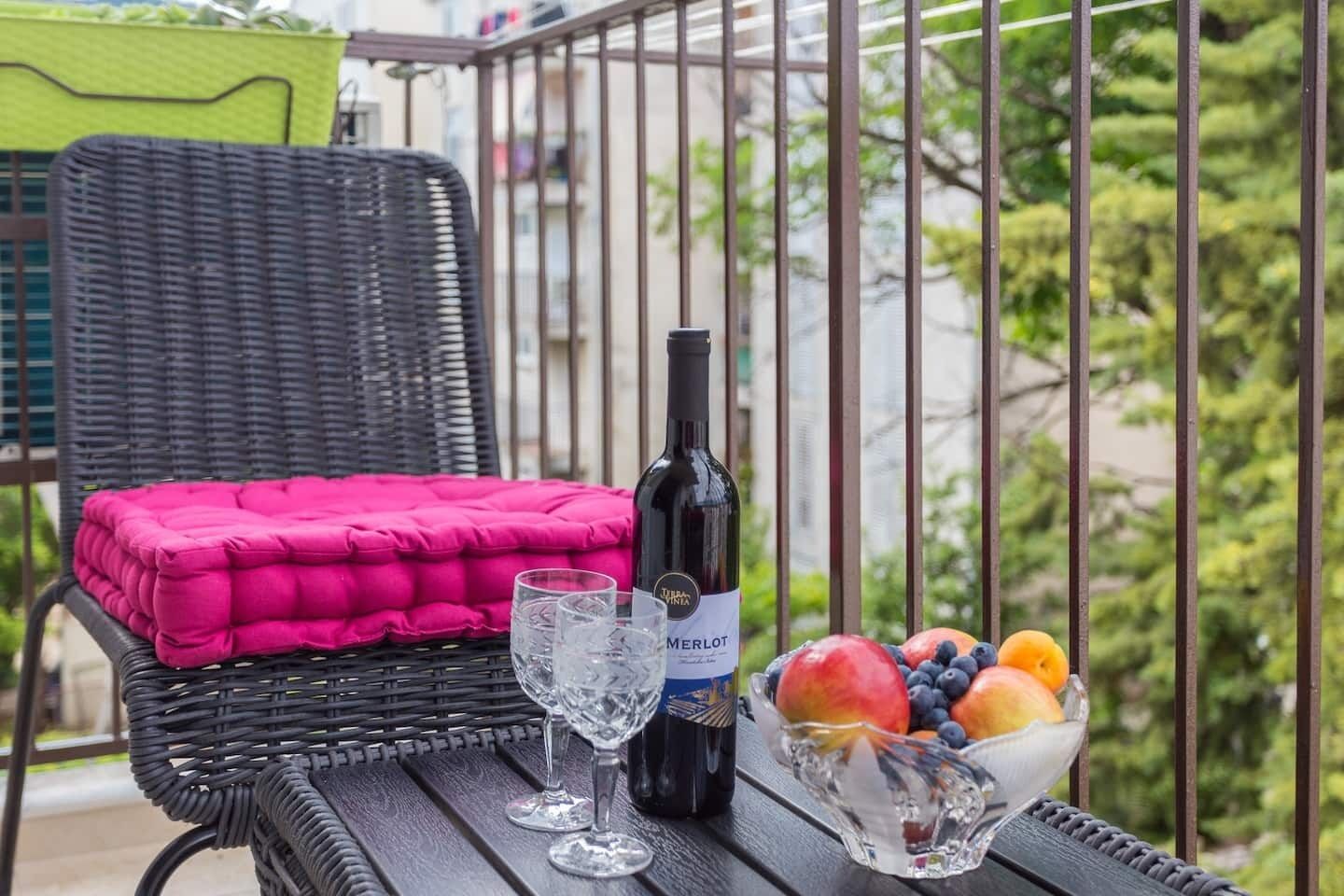 Un balcon avec un fauteuil en osier, une bouteille de vin Merlot, deux verres à vin et un saladier rempli de fruits.