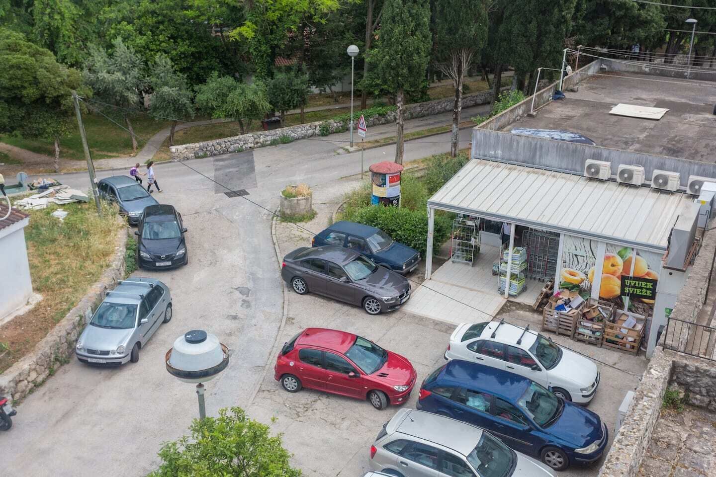 An aerial view of a parking lot with several cars, a small building with a sign, and a street with pedestrians.