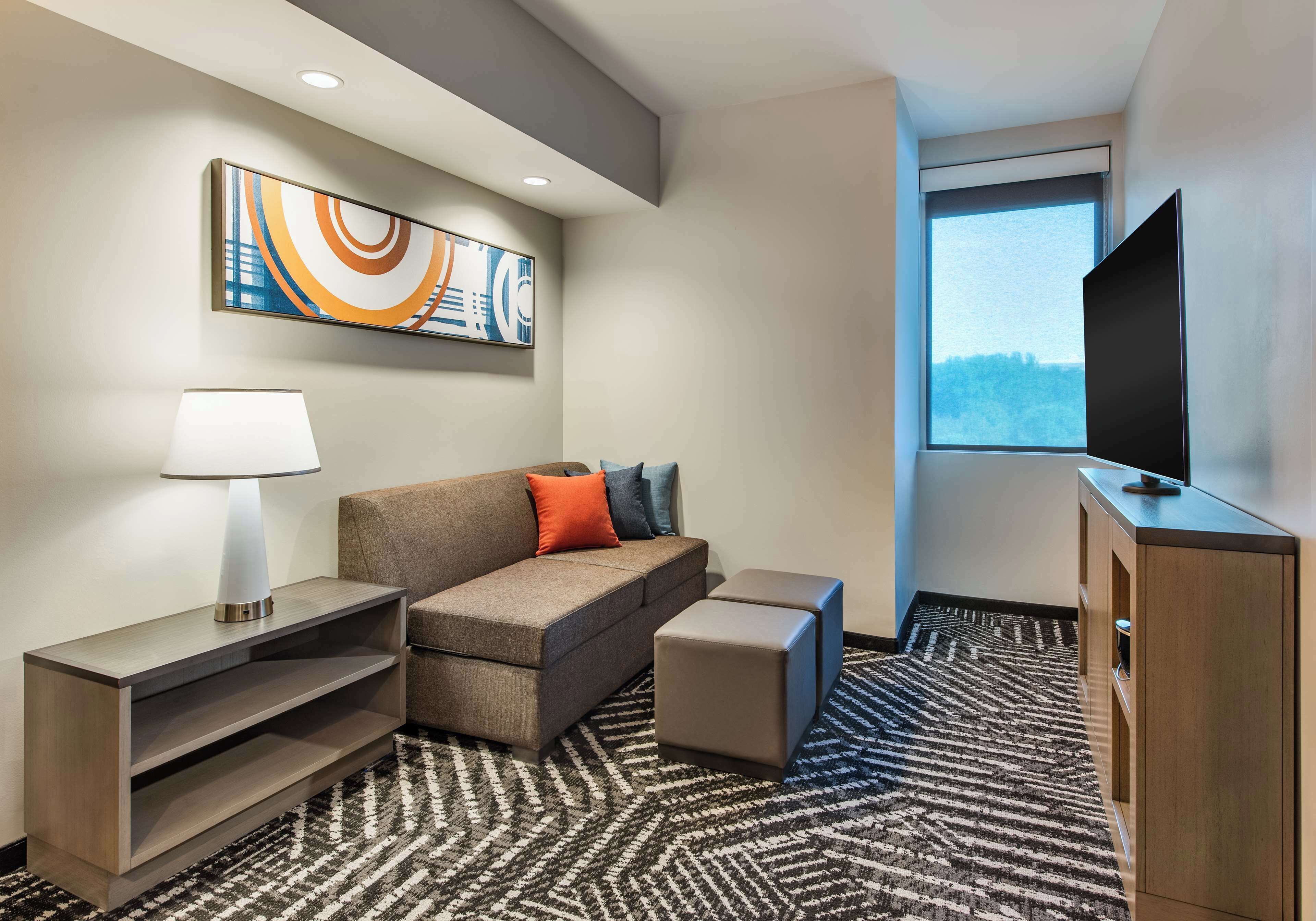 Una habitación de hotel moderna con sofá, reposapiés, televisión, lámpara y una obra de arte en la pared.