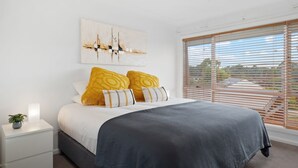 A bedroom with a bed, a nightstand, a potted plant, and a window with blinds. - Allinga Lodge, bright, beautiful and centrally located (Jindabyne)