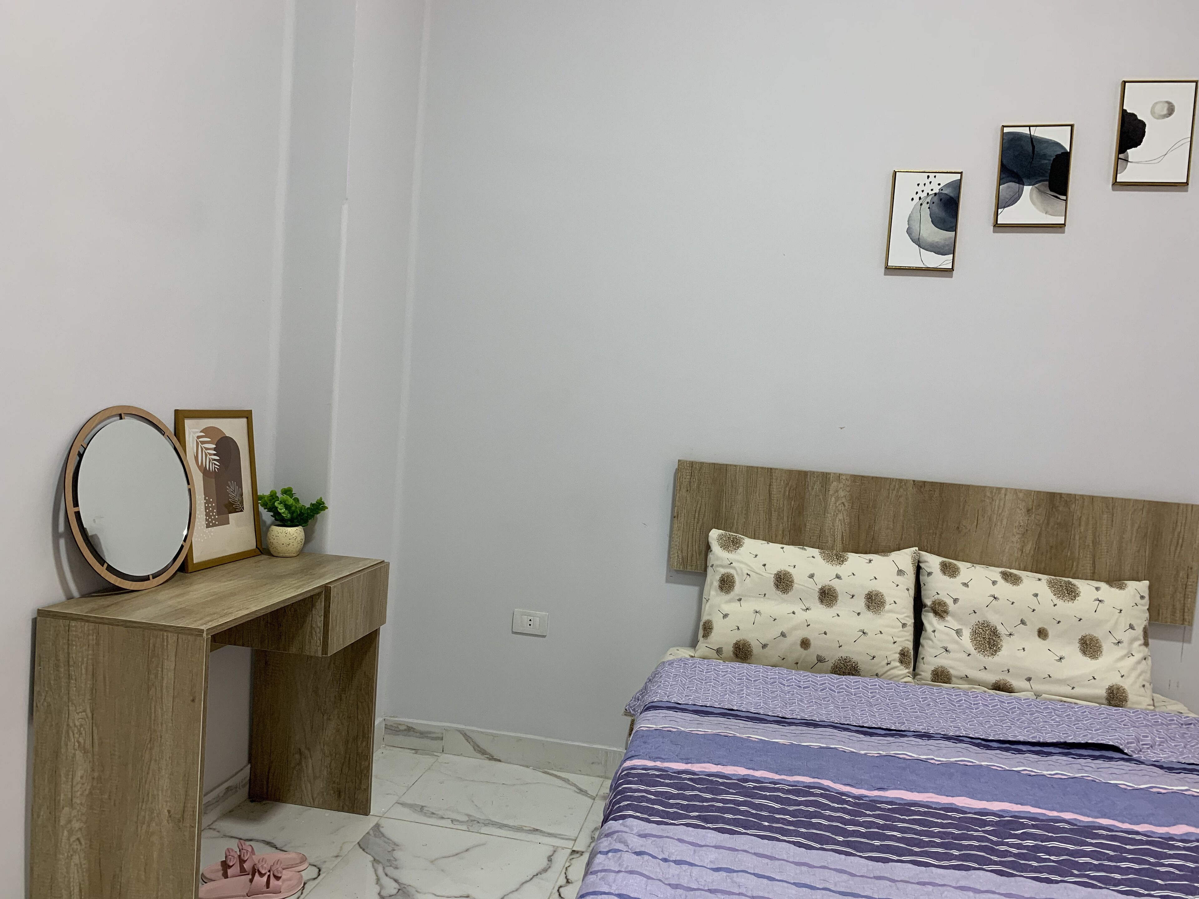 Une chambre à coucher avec une tête de lit en bois, un lit agrémenté d’oreillers à motifs, une table de chevet avec un miroir et des œuvres d’art encadrées accrochées au mur.