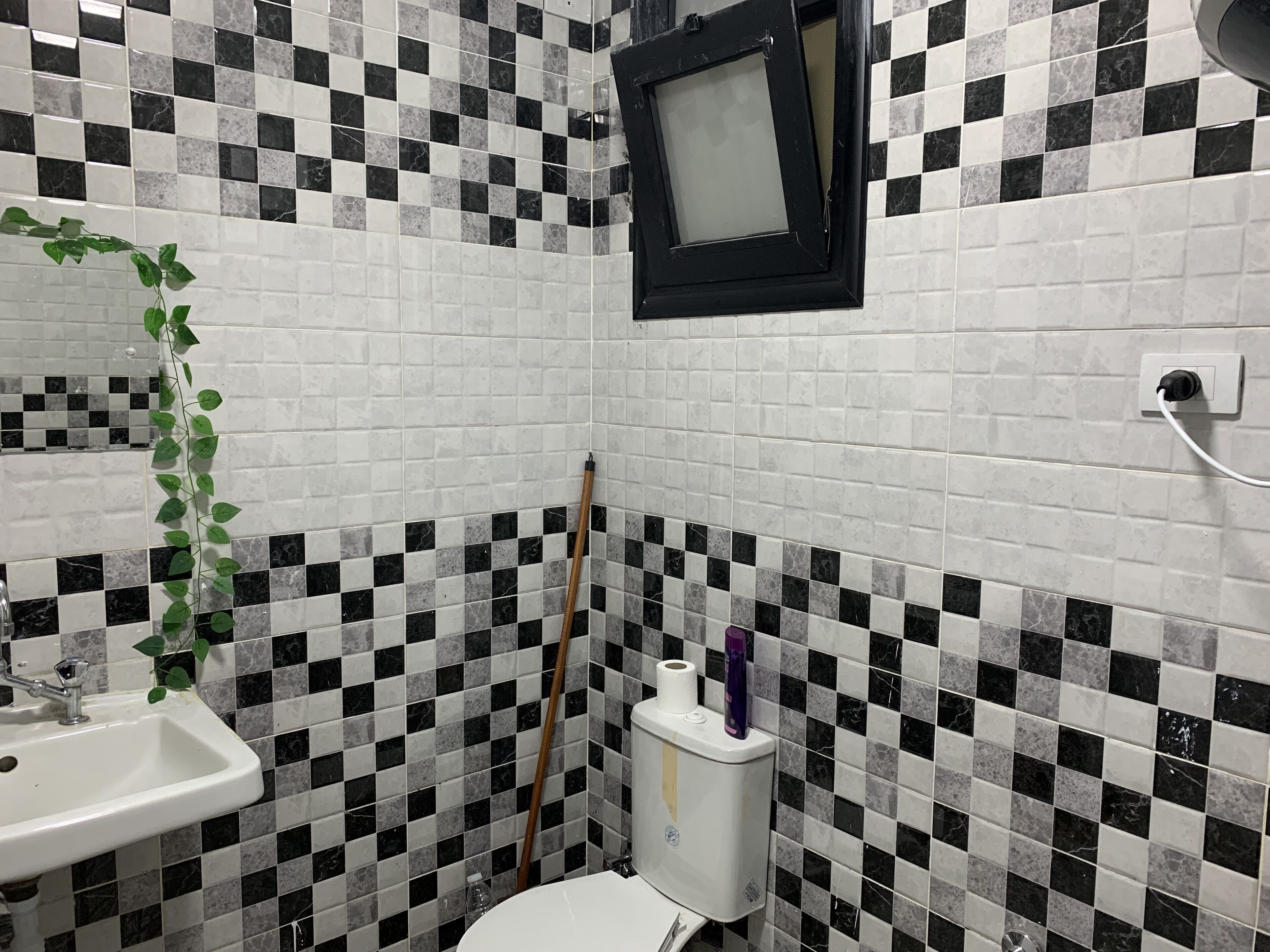 A bathroom with a checkered tile pattern, a white sink, a toilet, a broom, and a small window.